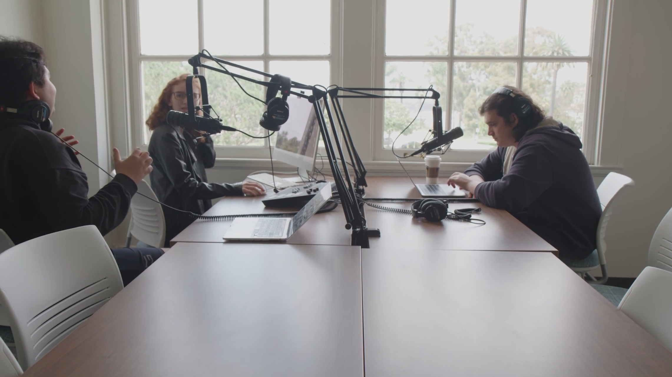 Three people sitting around a rectangular table speaking into podcasting speakers